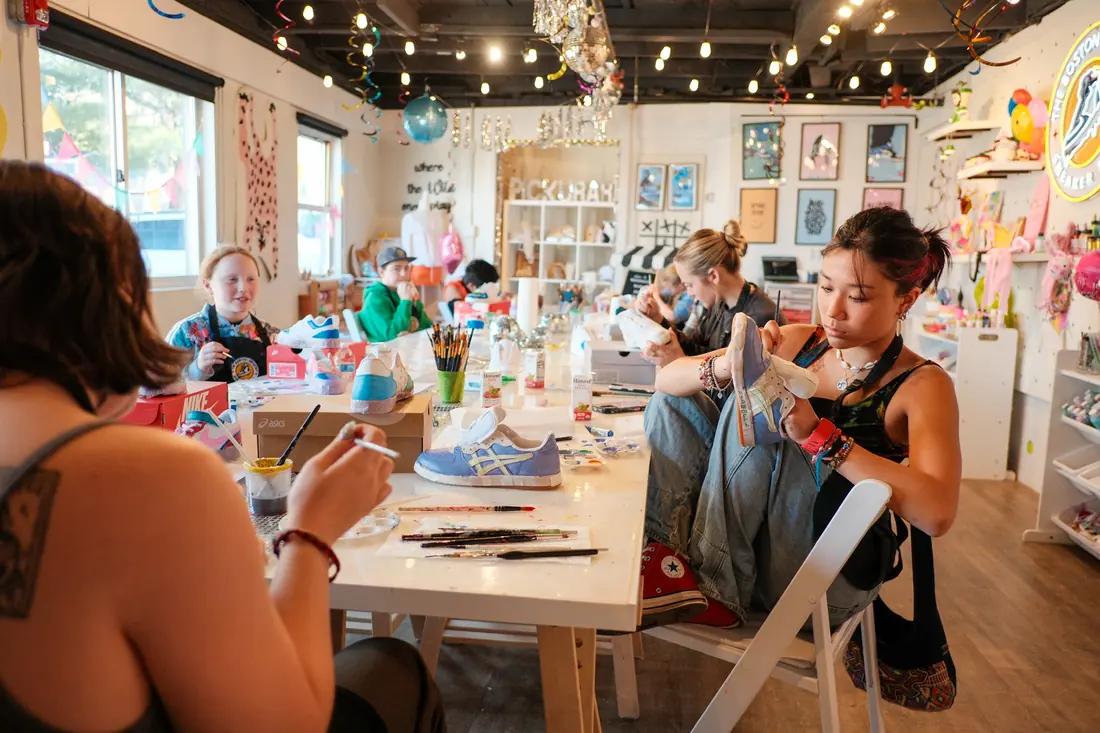 A teenager sits with her feet up painting a sneaker at a long studio table where others paint sneakers too