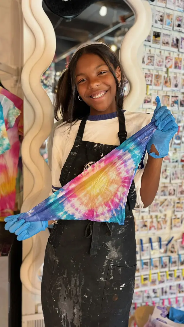 A smiling girl in a splattered apron and blue gloves holds up a spiral tie dye cloth in the studio