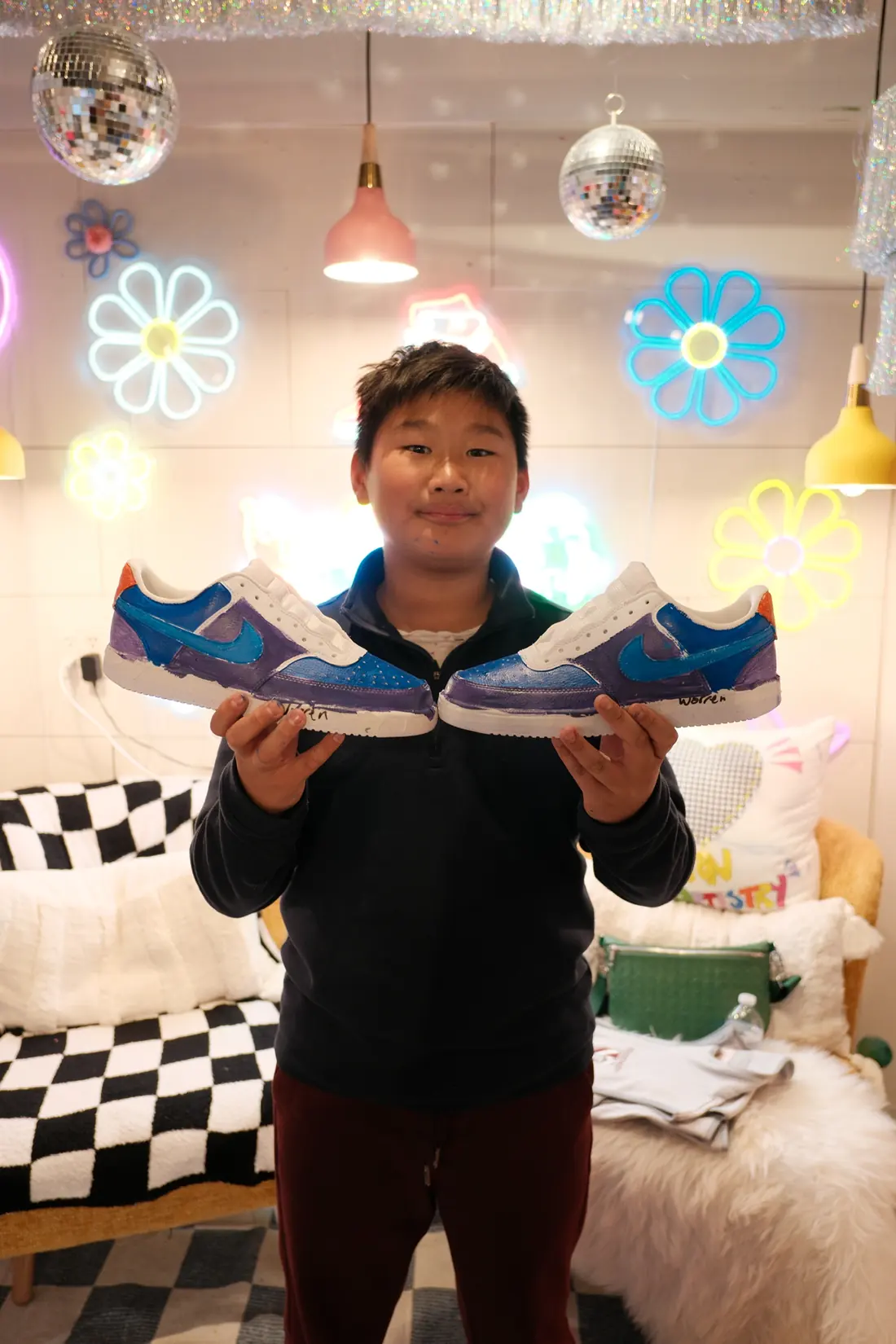 A boy in a navy pullover holds up a pair of sneakers painted blue and purple in front of neon flower lights